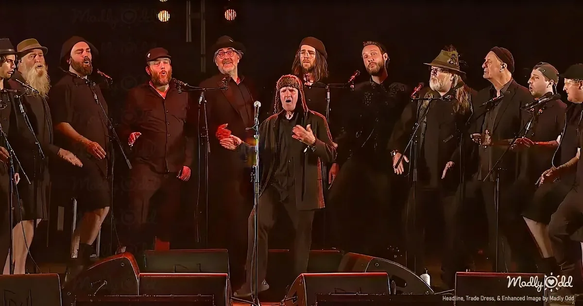 A man in a black coat and hat leads a choir of men in black attire on stage.