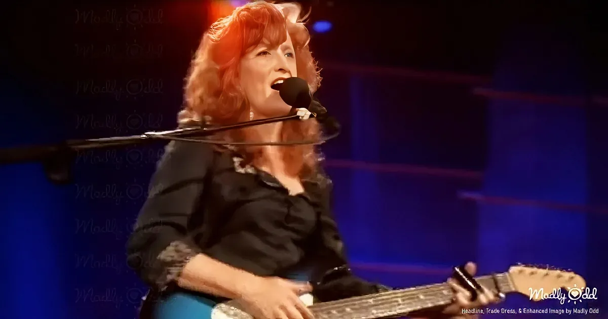 A woman with red hair sings into a microphone while playing a guitar on stage.