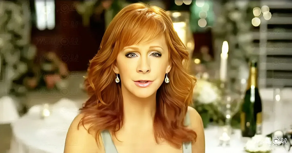A woman with long, wavy red hair and earrings, wearing a light top, stands in front of a festive table with champagne...