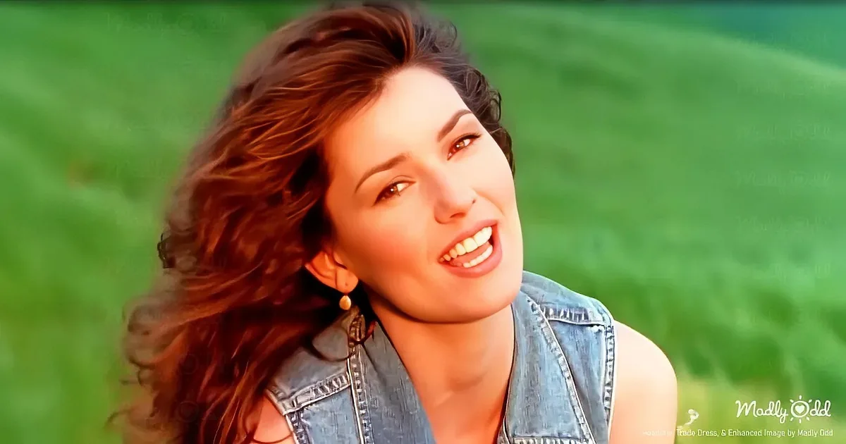 A woman with long, wavy brown hair and a denim vest smiles against a green, blurred background.