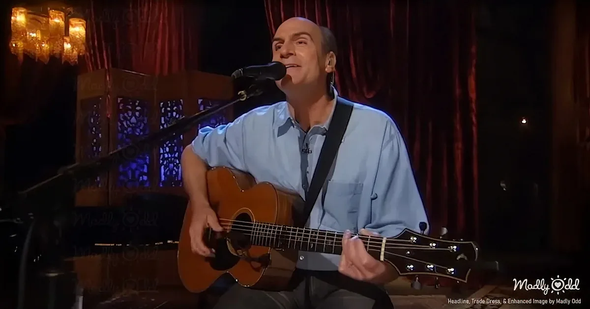 A man in a light blue shirt plays an acoustic guitar while singing into a microphone on stage.