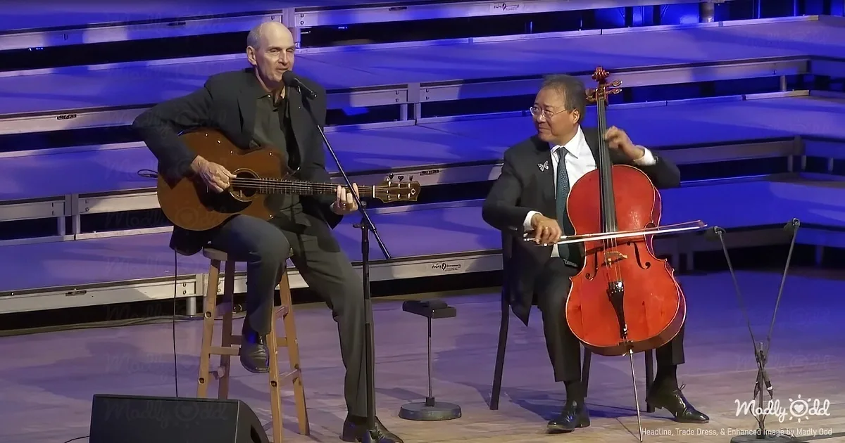 A man in a suit plays guitar while another in a suit plays cello on stage.
