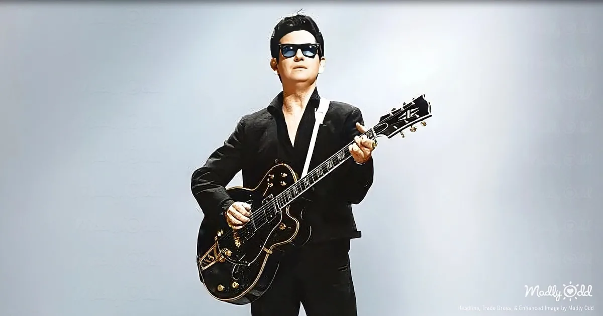 A man in a black suit and sunglasses holds a black electric guitar, standing against a light background.