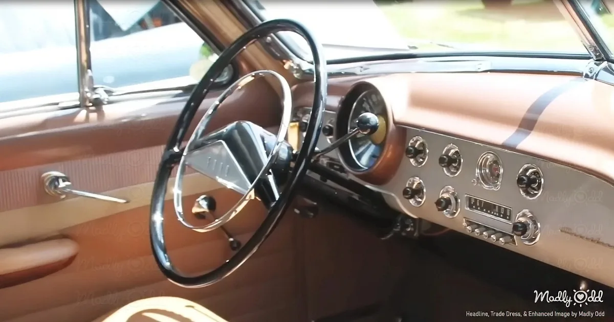 A vintage car's interior with a black steering wheel, tan dashboard, and chrome accents.