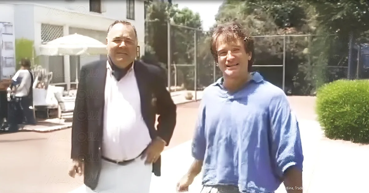 Two men stand outdoors, one in a suit, the other in casual attire, under bright sunlight.