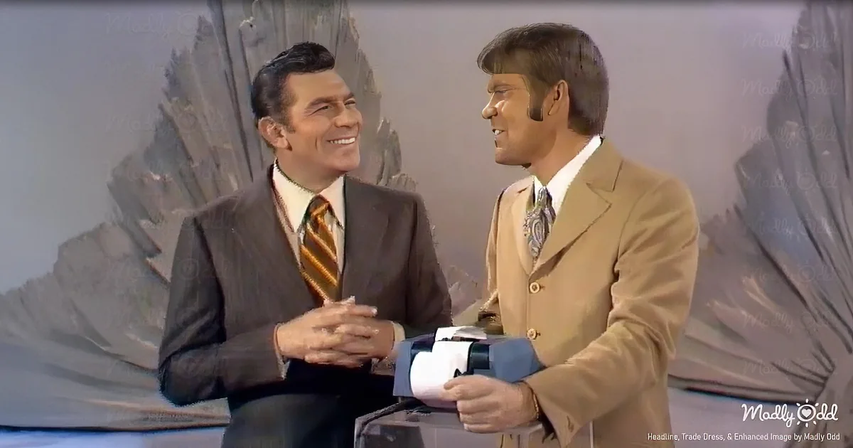 Two men in suits smile and interact, one holding a microphone, against a backdrop resembling a desert landscape.