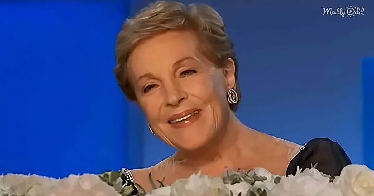 A woman with short gray hair, wearing a black dress and earrings, smiles while holding a bouquet of white flowers.