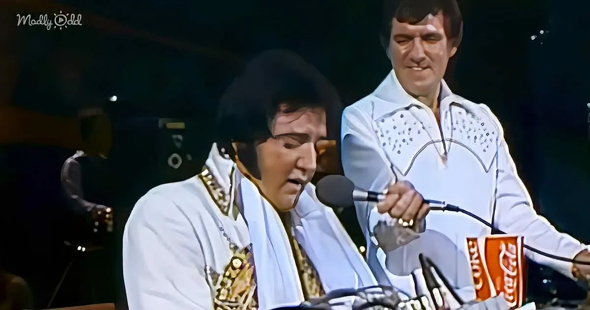 Elvis Presley in white stage outfit singing into microphone, another man in similar attire standing nearby.