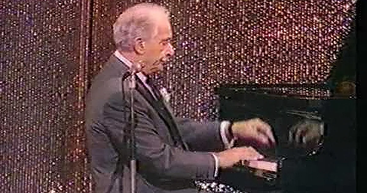 A man in a suit smiles while playing a piano on stage with a glittery backdrop.