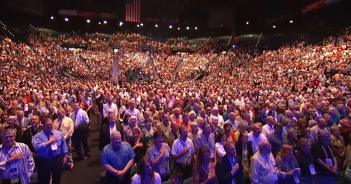 A large crowd of people in a stadium, many with hands over hearts, possibly during a patriotic event.