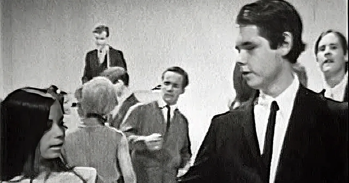 A man in a suit and a woman in a white dress are standing in a crowd, seemingly engaged in conversation.