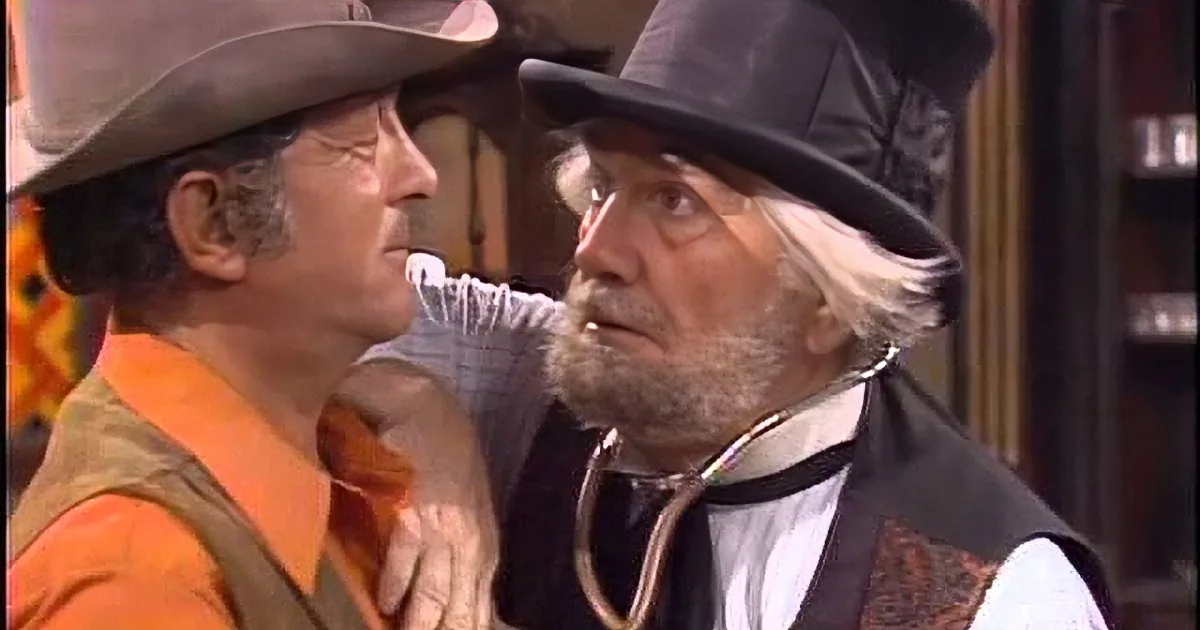 Two men in vintage attire, one in a cowboy hat, both smiling and holding a prop gun, in a staged scene.