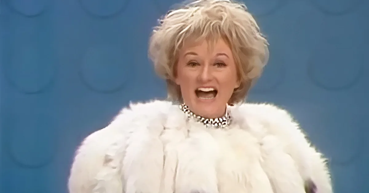 A person in a white fur coat and pearl necklace is smiling against a blue background.