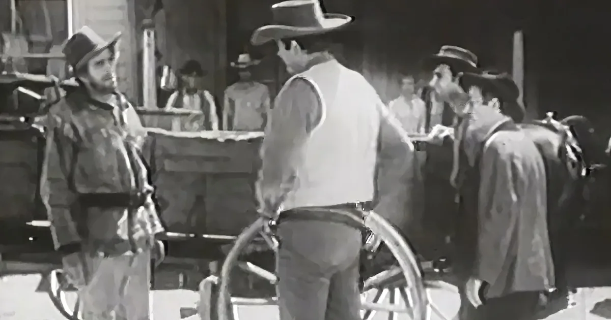 A man in a cowboy hat and vest stands beside a wheelbarrow, surrounded by others in Western attire.