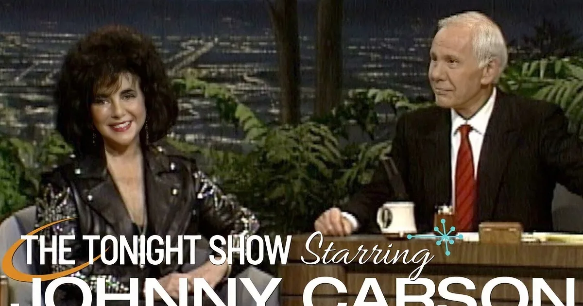 Woman in black leather jacket laughs with man in suit on talk show set.