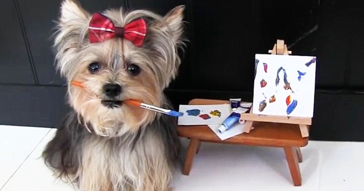 A fluffy dog with a red bow holds a paintbrush, standing next to an easel with a colorful painting.