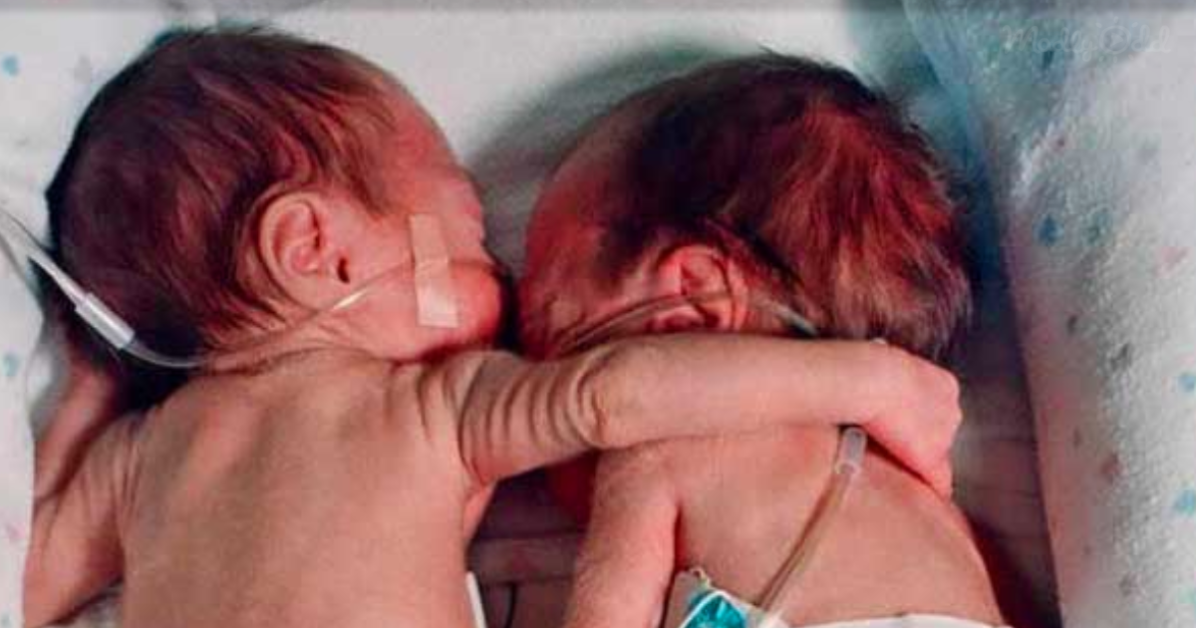 Two newborn babies, one with a nasal cannula, are lying close together, appearing to hug each other.
