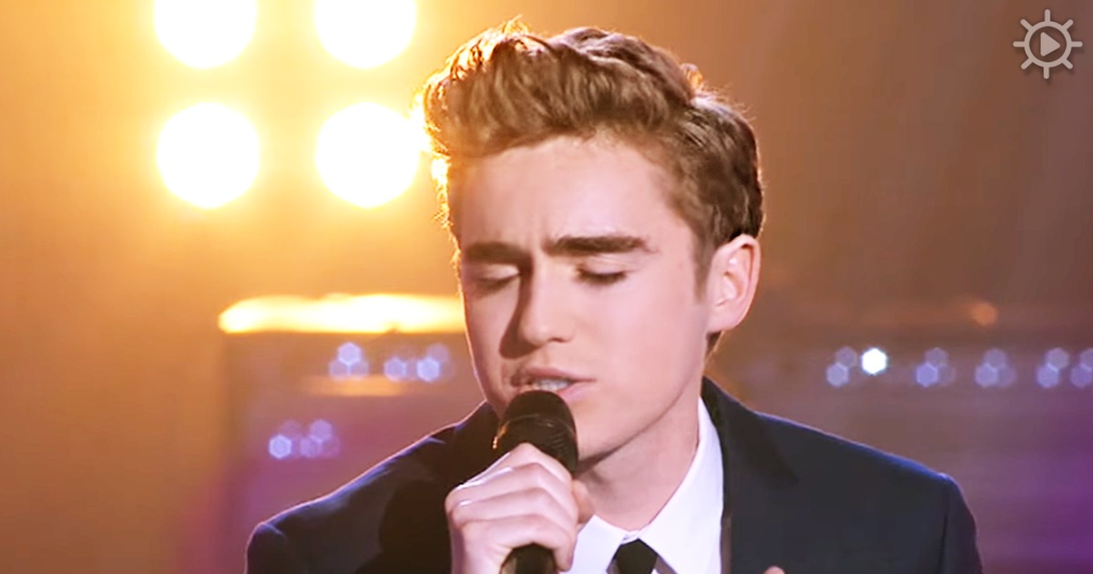 A young man in a suit sings into a microphone on a stage with bright lights.