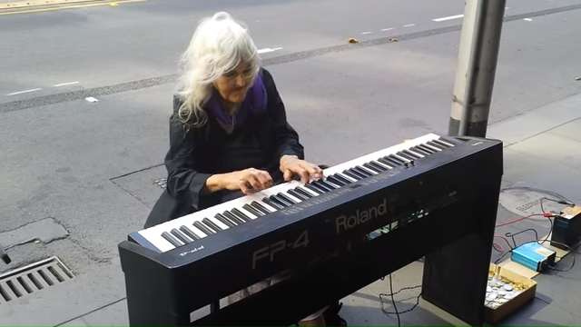An elderly woman with white hair plays a Roland keyboard on a city street.
