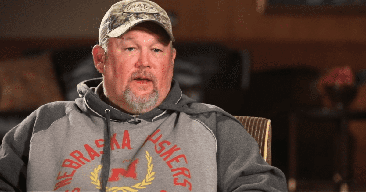 A man wearing a camouflage cap and a Nebraska Cornhuskers hoodie sits in a chair, speaking.