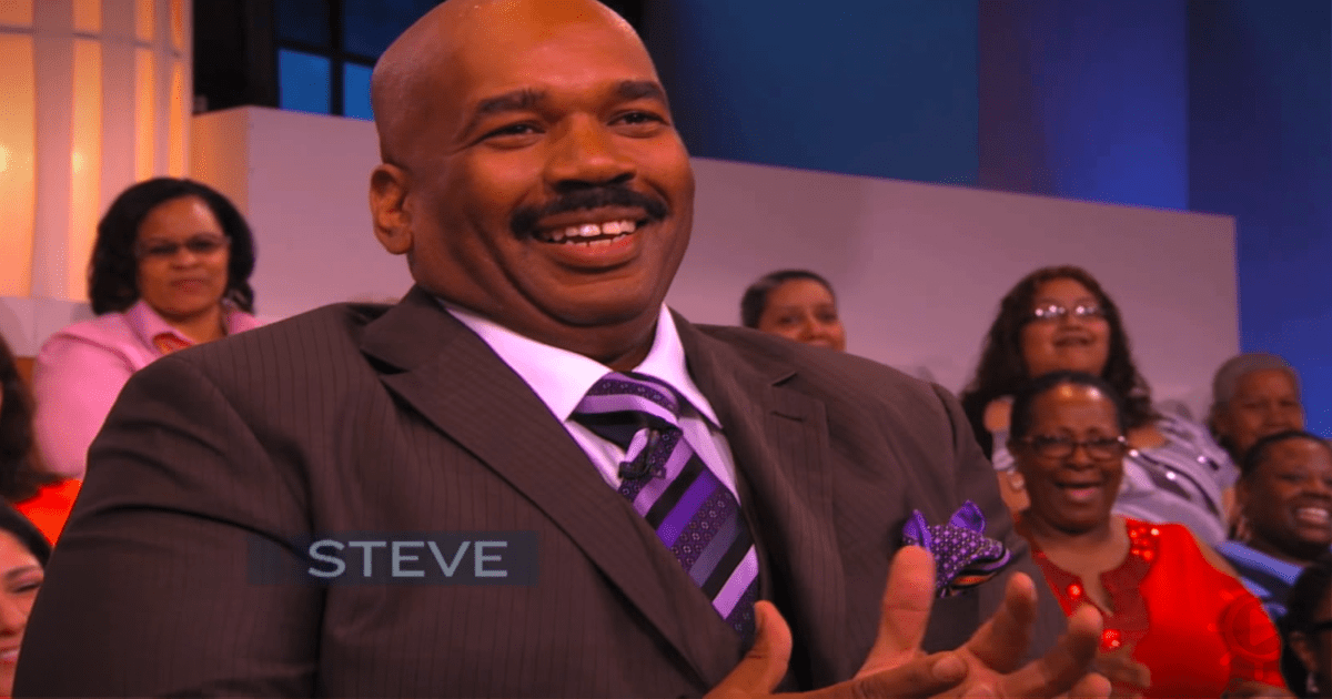 A man in a suit smiles, gesturing with his hands, with a group of people in the background.