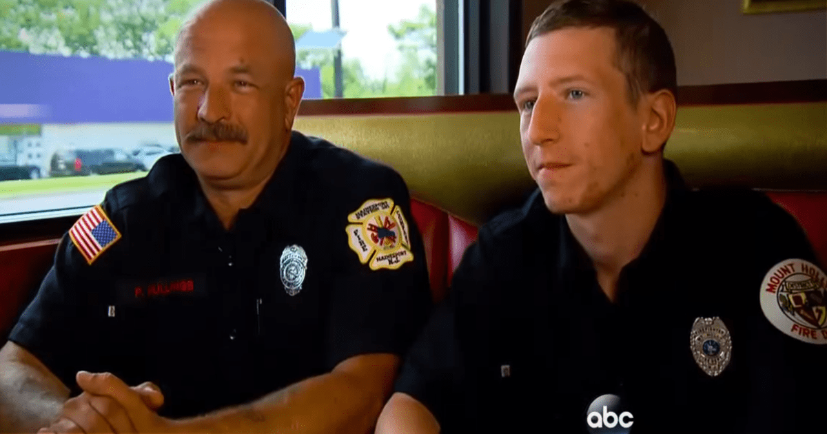 Two firefighters in uniform sit side by side, one with a mustache, both with patches and badges on their shirts.