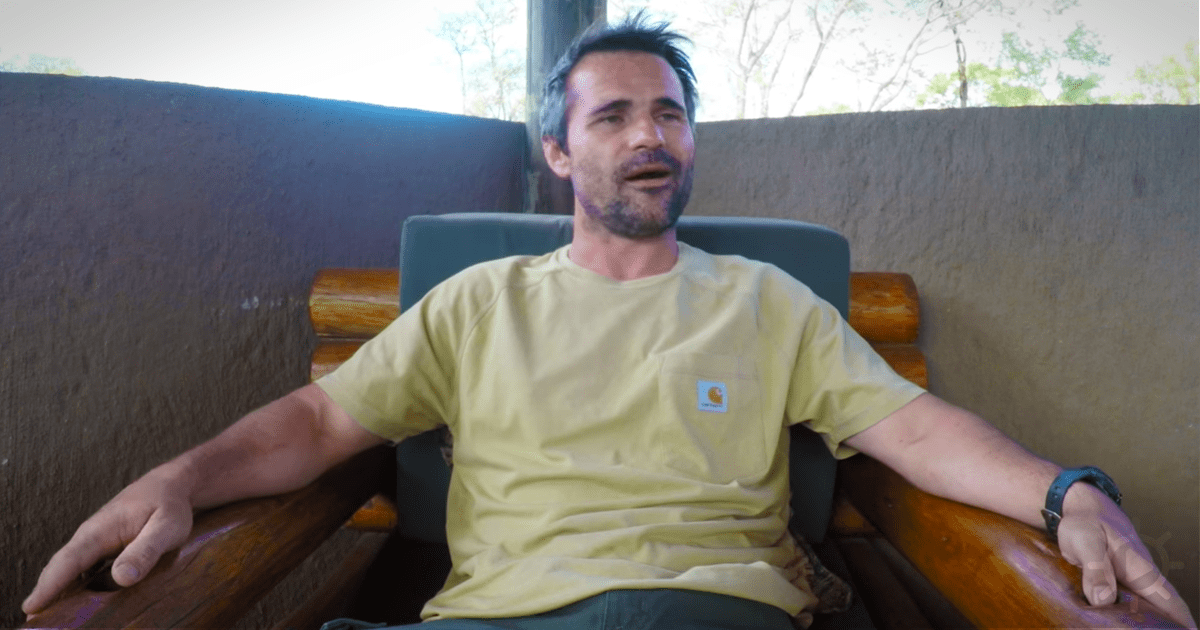 A man in a yellow Carhartt shirt sits on a wooden bench, looking to the side with a relaxed posture.