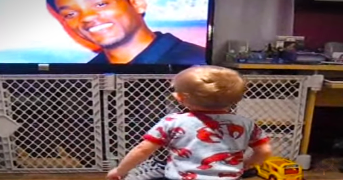 A baby in pajamas sits in front of a TV, watching a man's face on the screen.