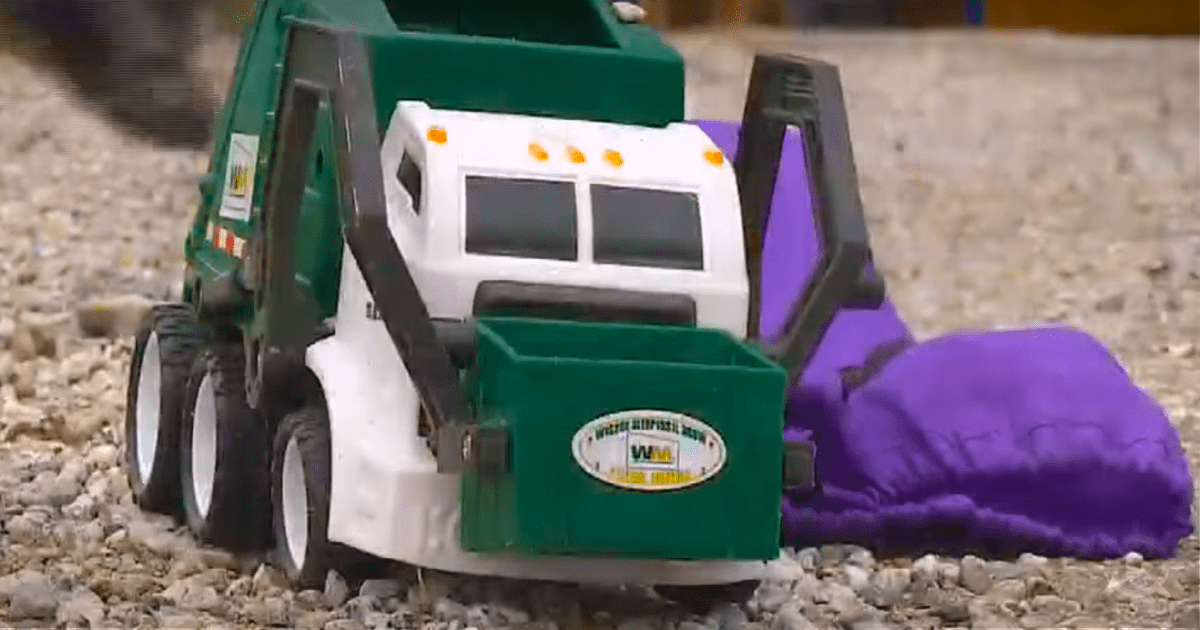 A green and white toy garbage truck is on a gravel surface, with a purple object nearby.