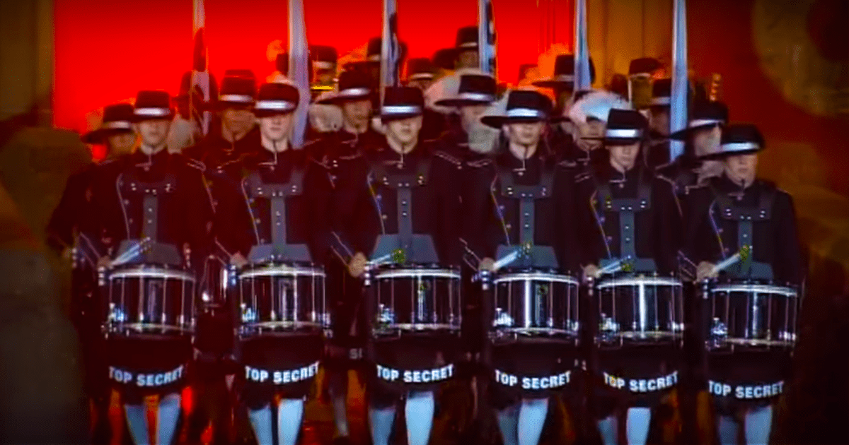 A group of uniformed drummers in black uniforms with "TOP SECRET" written on their pants march in formation.