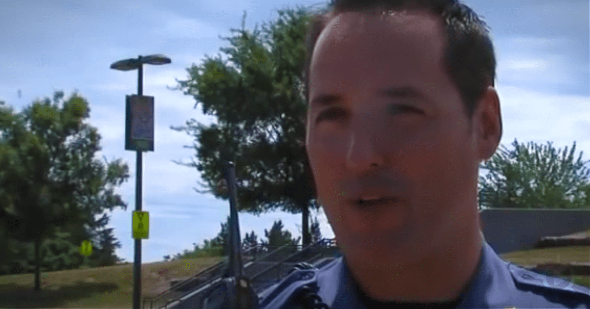 A man in a uniform is speaking outdoors, with trees and a sign in the background.