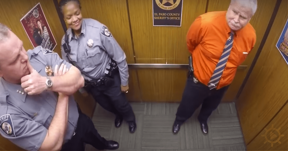 A man in an orange shirt is being restrained by two sheriff's deputies in an office setting.