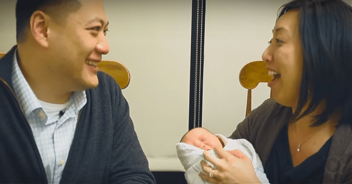 A man and woman are laughing while holding a baby wrapped in a blanket.