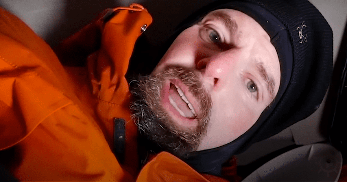 A man with a beard and a black hooded jacket is looking up, possibly in a vehicle.