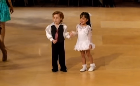Two children, a boy in a vest and a girl in a white dress, stand on a dance floor, smiling and holding hands.