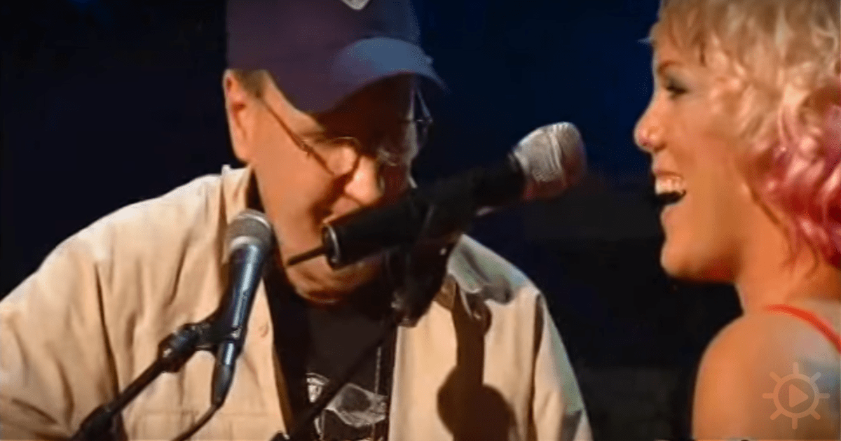 A man in a cap and glasses holds a microphone, singing with a woman smiling beside him.