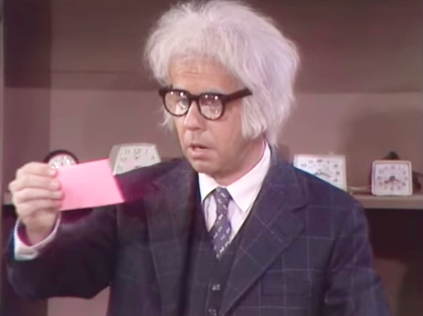 A man in a suit and glasses holds a pink card, appearing surprised or confused.