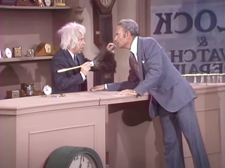 A man in a suit leans over a desk, interacting with another man in a suit, who is holding a pencil.