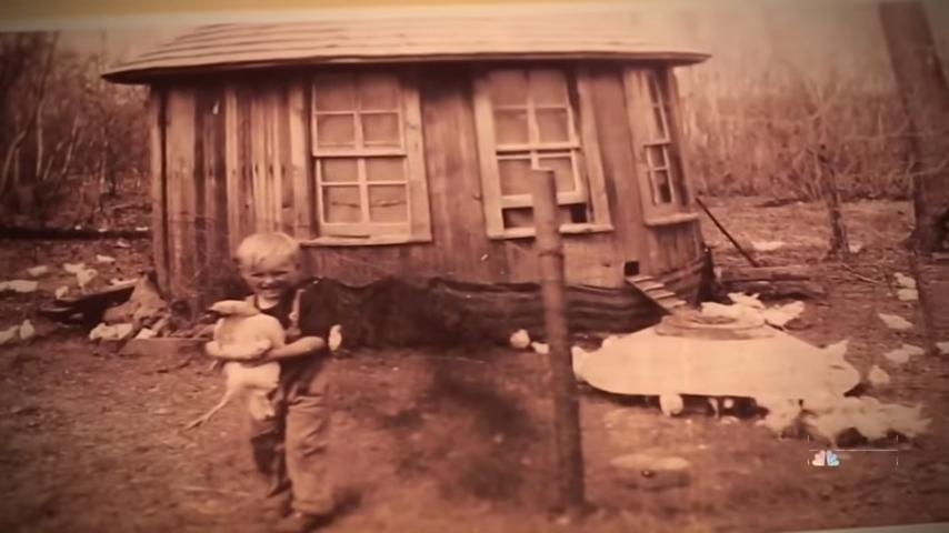 A young boy stands in front of a rustic wooden shed, surrounded by chickens and a pile of white feathers.