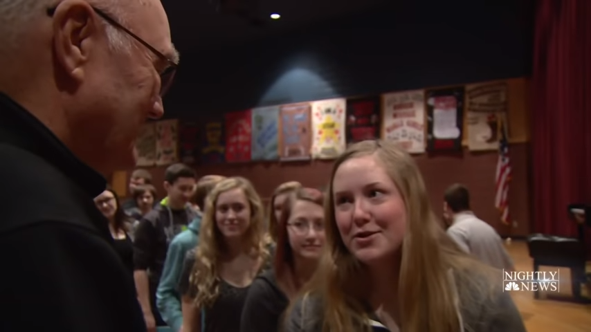 A man in glasses speaks to a young woman in a school setting with posters and an American flag in the background.