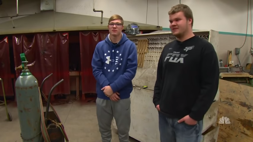 Two young men stand in a workshop, one in a blue hoodie, the other in a black sweatshirt, surrounded by tools and equipment.