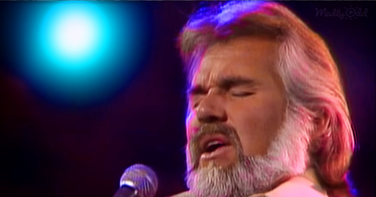 A man with long hair and a beard sings into a microphone under colorful stage lighting.