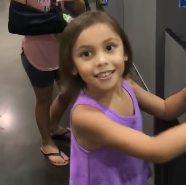 A young girl in a purple tank top is holding a phone to her ear, smiling, with a person in pink shorts and black sandals...