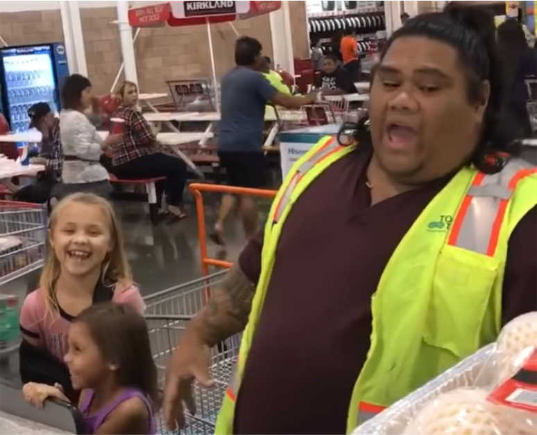 A person in a yellow safety vest holds groceries, smiling, with a child in the background.