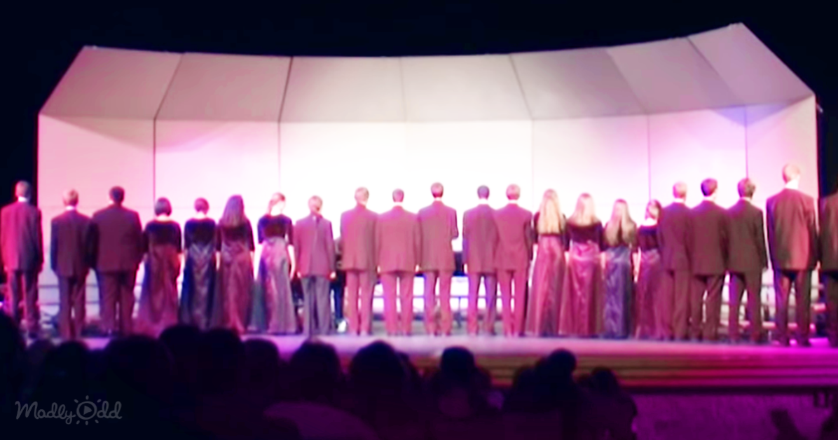 A choir stands in a line on stage, facing away from the audience, under a large white tent.
