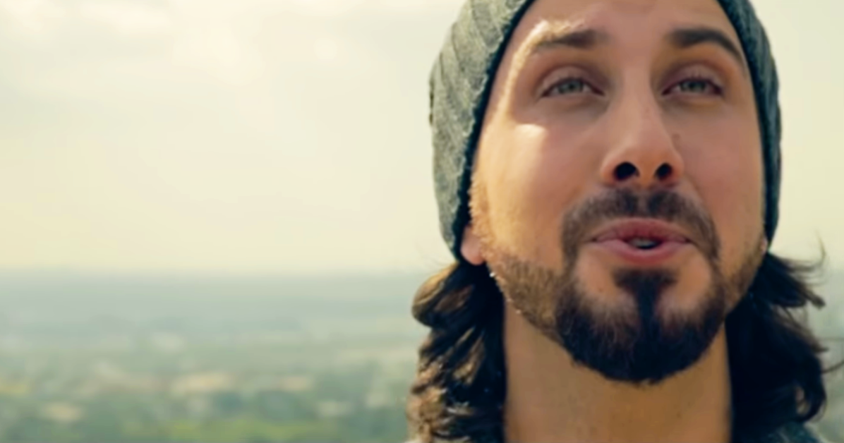 A man with long hair and a beard, wearing a beanie, looks upward with a serene expression against a blurred landscape.