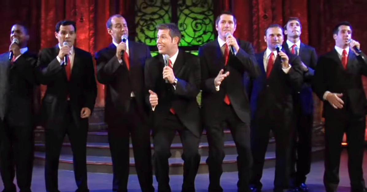 A group of men in suits sing on stage, holding microphones, with a red and green backdrop.