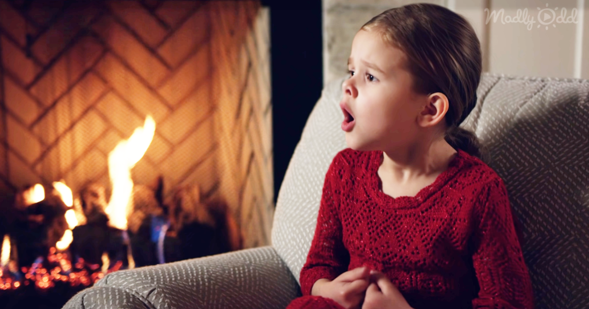 A young girl in a red sweater sits on a couch, mouth open, gazing at a fireplace with dancing flames.