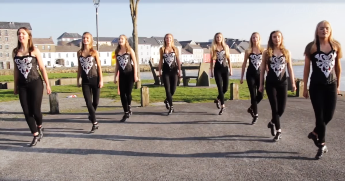 Eight dancers in black and white costumes perform a synchronized step routine on a paved path near a waterfront.