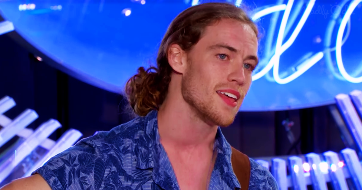 A man with long hair in a ponytail, wearing a blue floral shirt, is speaking in front of a blue-lit background with neon...